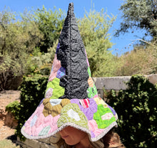 Load image into Gallery viewer, A person outdoors wearing a tall, pointed witch-style quilted hat with a wide brim. The hat features a black polka-dot pointed top and a multicolored patchwork design with pink, green, purple, yellow, and white sections.