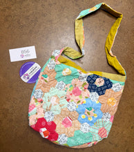 Load image into Gallery viewer, A handmade quilted shoulder bag with a multicolor patchwork design featuring hexagonal fabric pieces in pink, blue, green, and yellow, with a solid mustard yellow strap. It is displayed on a brown textured surface next to a white paper tag and a purple circular sticker.